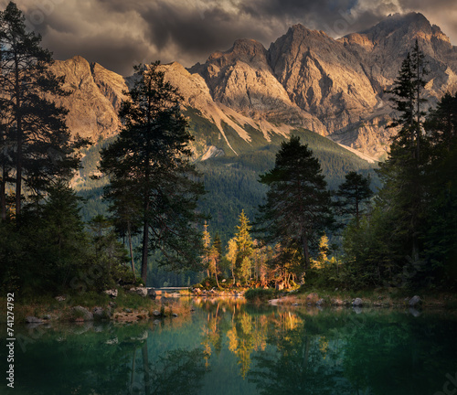 Dramatic scenery with a mountain lake landscape before sunset, with dark clouds and a sunlit little island framed by tree silhouettes and reflected in the teal water