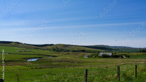 Expansive rural landscape with a winding stream, green fields, and a blue sky, evoking a sense of tranquility.