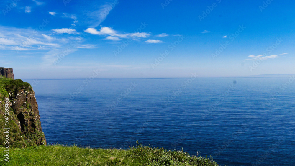 A coastal landscape with towering cliffs overlooking a calm blue sea under a bright blue sky, with greenery in the foreground.
