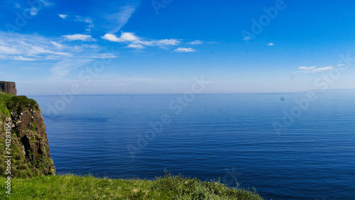 A coastal landscape with towering cliffs overlooking a calm blue sea under a bright blue sky, with greenery in the foreground.