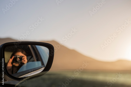 Chica sacándose un retrato en el reflejo del espejo retrovisor del coche durante las vacaciones, con una cámara de fotos