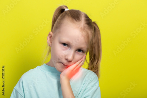 A beautiful little girl of seven years old holds her red, inflamed cheek with her hand. Concept of tooth pain in children, caries, gum inflammation and gingivitis. Yellow background photo