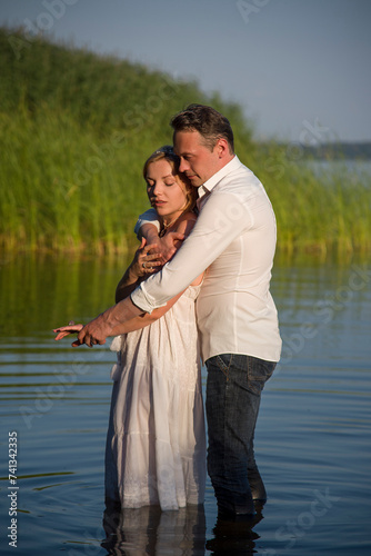 beautiful happy hugging couple, bride and groom in white dresses walking in forest river on sunny summer day and holding each other hand by hands