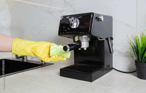 A girl s hand in a yellow glove washes a black coffee machine with a sponge with foam and cleaning agent in the kitchen against a background of a white wall with porcelain tiles. photo