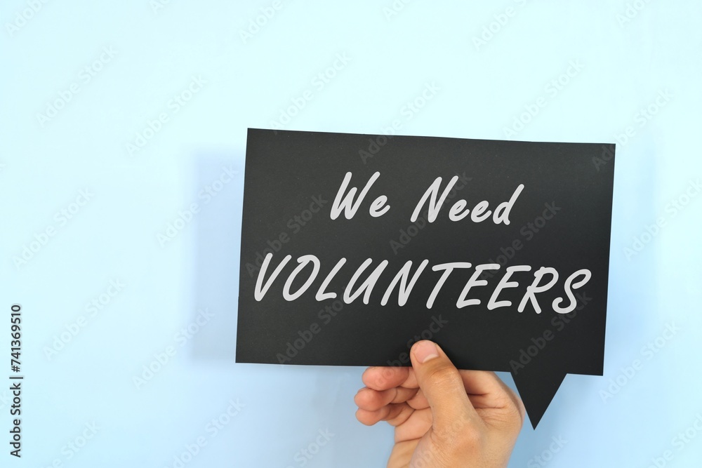 Human hand holding black speech bubble signage with written phrase We Need Volunteers. Volunteering concept.