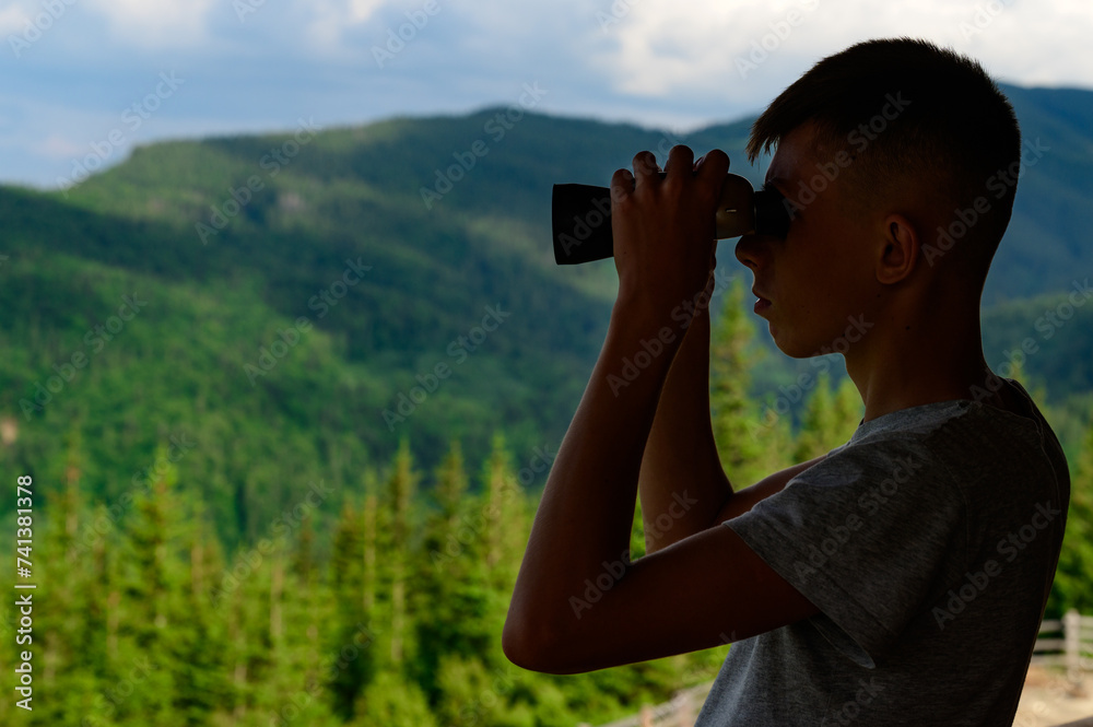 Boy teenager looks through binoculars at the mountains.