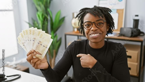 Joyful young black woman with dreadlocks, showcasing danish krone banknotes at the office, smiling and pointing confidently! photo