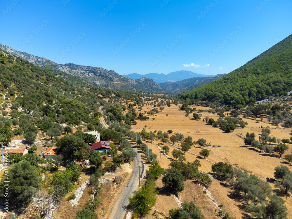 Drone view of a rural road in a gorge of mountains with buildings, houses and trees in Turkey. Agricultural land at the foot of the mountains. Hot sunny day, romance of traveling by car on vacation.