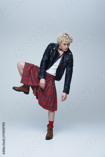 Fusion of cultures. Full length portrait of man in Scottish kilt and punk leather jacket posing against grey studio background. Concept of fusion, art, style, queer, uniqueness, self-expression. Ad