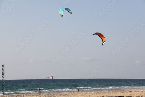 people flying their colorful kites at the beach while in the far distance a cargo ship sails by
