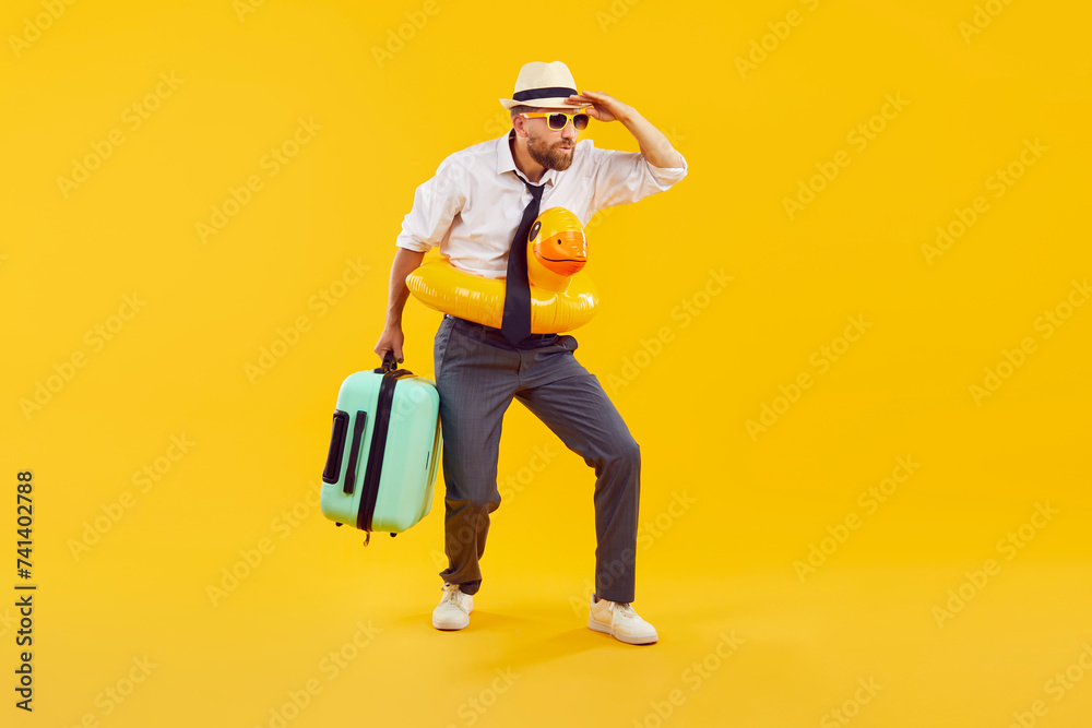 Business man with summer holiday travel bag, in funny orange inflatable swim lifebuoy standing on yellow background, holding hand above eyes, looking in distance, searching where to have fun and relax Business man with summer holiday travel bag, in funny orange inflatable swim lifebuoy standing on yellow background, holding hand above eyes, looking in distance, searching where to have fun and relax