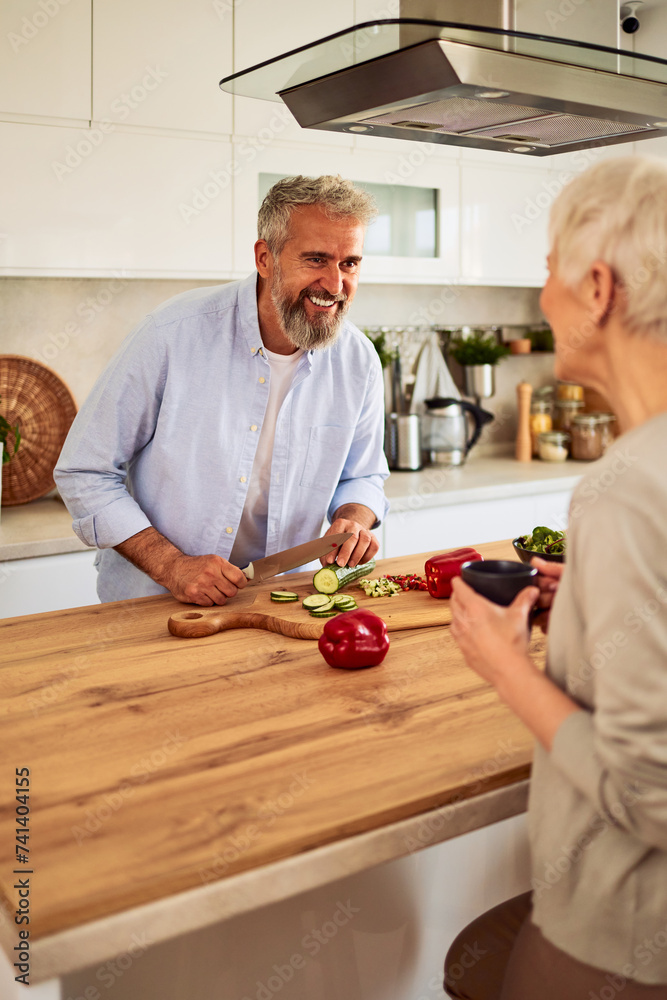 A cheerful senior adult man laughing with his wife in the kitchen while cutting vegetables for a salad A cheerful senior adult man laughing with his wife in the kitchen while cutting vegetables for a salad