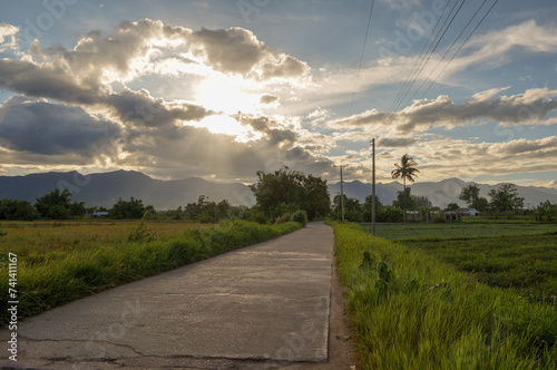 The road in the middle of the field in the evening saw clouds blocking the sun and mountains in Thailand. .