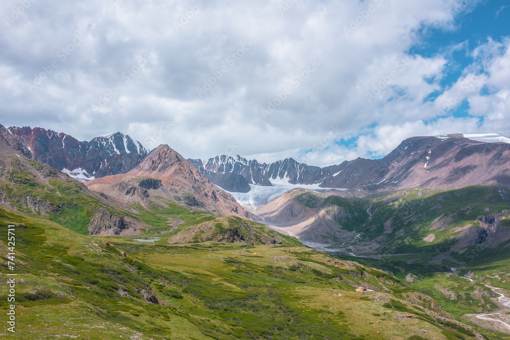 Colorful vast scenery of wide green alpine valley with panoramic view to large snow-capped top, sharp rockies, rocky pointy peak, snowy mountain range and big glacier tongue far away under cloudy sky.