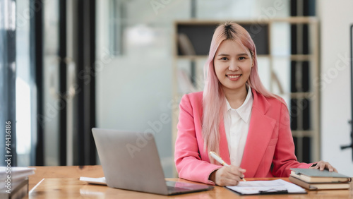 Confident young asian businesswoman with pink hair working on laptop in office. Professional attire and modern workplace concept.