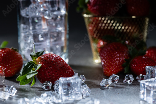 Strawberries nestled amidst ice cubes with a drink in the background against a dark backdrop