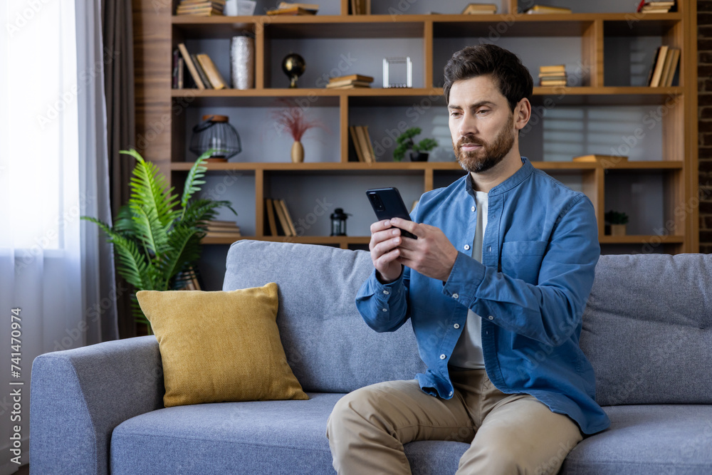Focused bearded man using smartphone while sitting comfortably on a grey couch at home