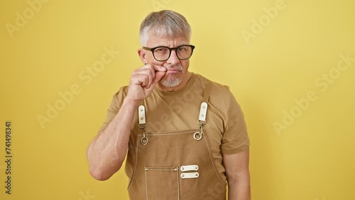 Shush! middle-aged, grey-haired man in glasses and apron gives a silent, secret zip gesture, hallmark of confidentiality. he's standing isolated against a yellow background, lips firmly shut. photo