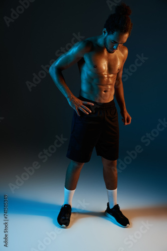 young shirtless african american man in black shorts posing in gym on dark lighted background