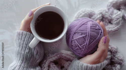 Female hands holding a cup of black coffee and a ball of purple yarn on a white table
