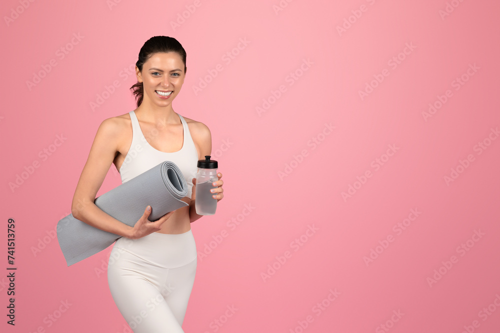 A smiling, fit woman in a white tank top and leggings holds a yoga mat under one arm and a water