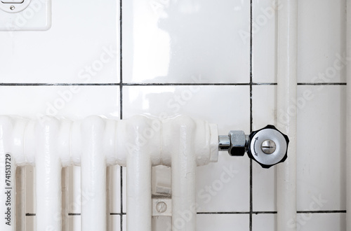 Cast iron home radiator thermostatic control valve. Front view, white ceramic bathroom tile wall, no people photo