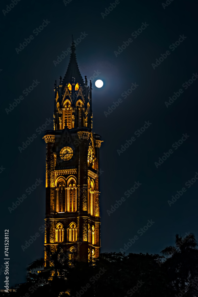 Night illuminated, UNESCO heritage building of Rajabai Tower , Mumbai with full moon in the background sky.