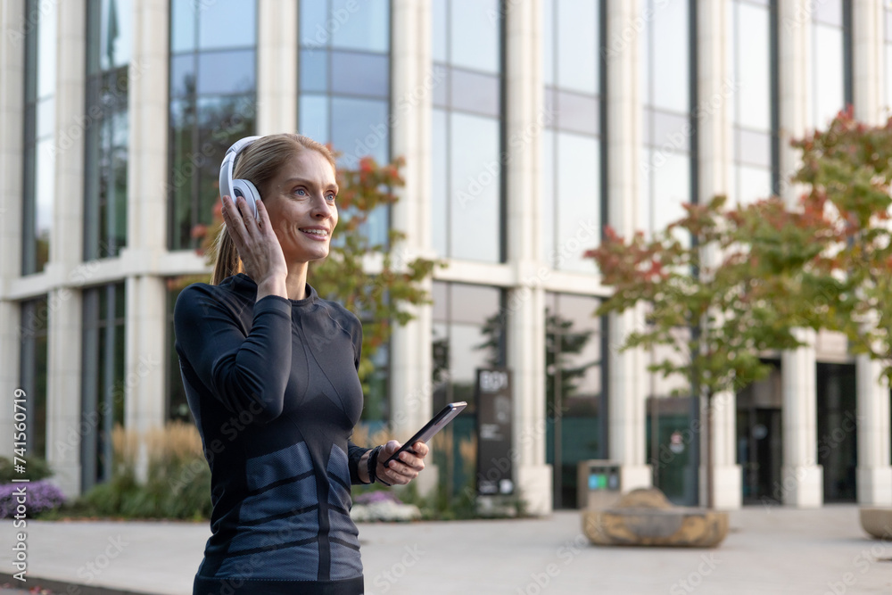 Smiling active woman with headphones enjoying music on smartphone in a city