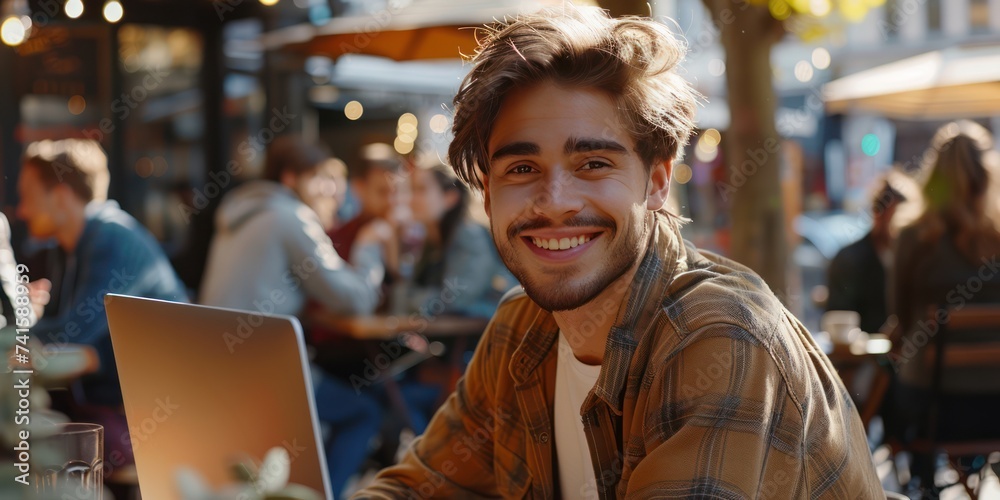 Young Male start-up entrepreneur, happy and smiling, working on a computer in the middle of a busy coffee shop with people in the background