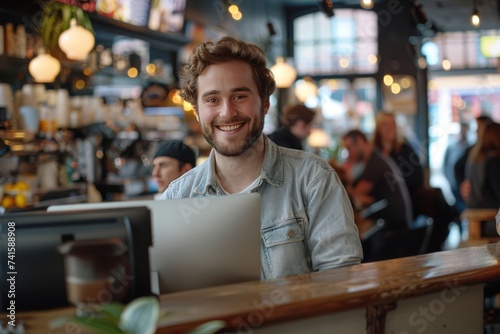 Young Male start-up entrepreneur, happy and smiling, working on a computer in the middle of a busy coffee shop with people in the background