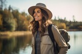Beautiful smiling young woman with backpack and hat standing near the lake.