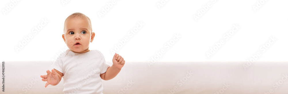 Portrait of beautiful little baby, adorable girl, toddler in white clothes posing on bed against white background. Concept of childhood, family, care, infancy. Banner. Empty space to insert text, ad