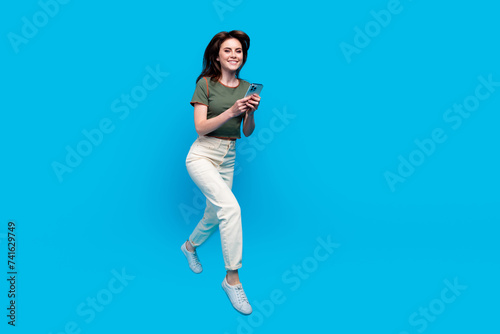 Full size photo of smart lovely woman wear green t-shirt run to empty space with smartphone in hands isolated on blue color background