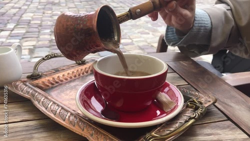Woman hand pouring greek coffee from a copper pot on a red cup placed on a tray in outdoor cafe