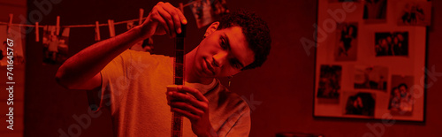 focused african american photographer examines developed film strip, bathed in red darkroom, banner