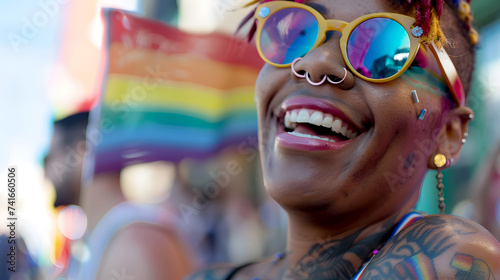 Non-binary person wearing sunglasses, smiling, celebrating gay pride day with rainbow flag