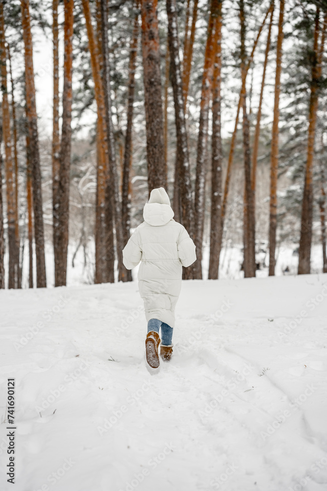 A cute young girl in a white jacket and hat runs and has fun in the winter in the forest, the girl has a good time outdoors, active lifestyle