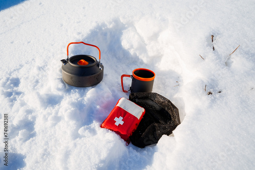 A red first aid kit complements the set of equipment for a winter hike in the mountains, a kettle is in the snow, a coffee mug, the equipment is in a snowdrift. © Aleksey