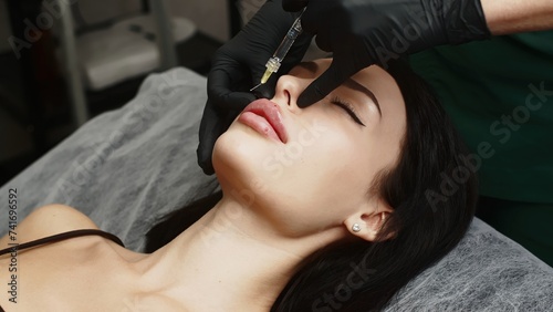 A surgeon wearing medical gloves carefully and slowly injects hyaluronic acid into a woman s lips using a syringe. lip augmentation procedure. beauty injections.