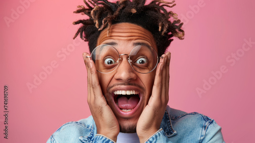 man with curly hair and glasses, his mouth open in a surprised expression, hands raised to the sides of his face against a pink background. © MP Studio