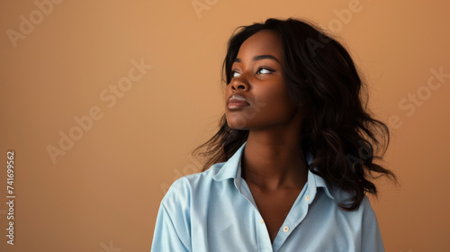 young African woman looking upwards, with a thoughtful expression, wearing a light blue shirt against a tan background.