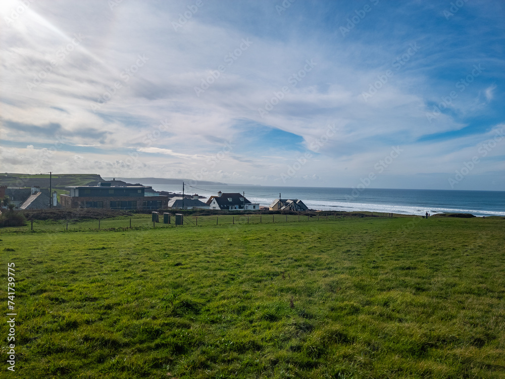 Coastal landscape with green fields, houses, and a distant beach under a blue sky with clouds.