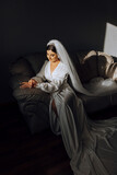 an incredibly beautiful bride in the morning in a white robe and a long veil sits on the bed in her bedroom. The bride poses in the morning before the wedding ceremony.