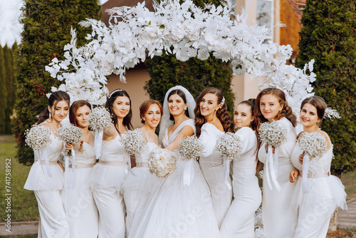 Wedding photography. The bride in a wedding dress and her friends in white dresses pose with bouquets near a white arch.