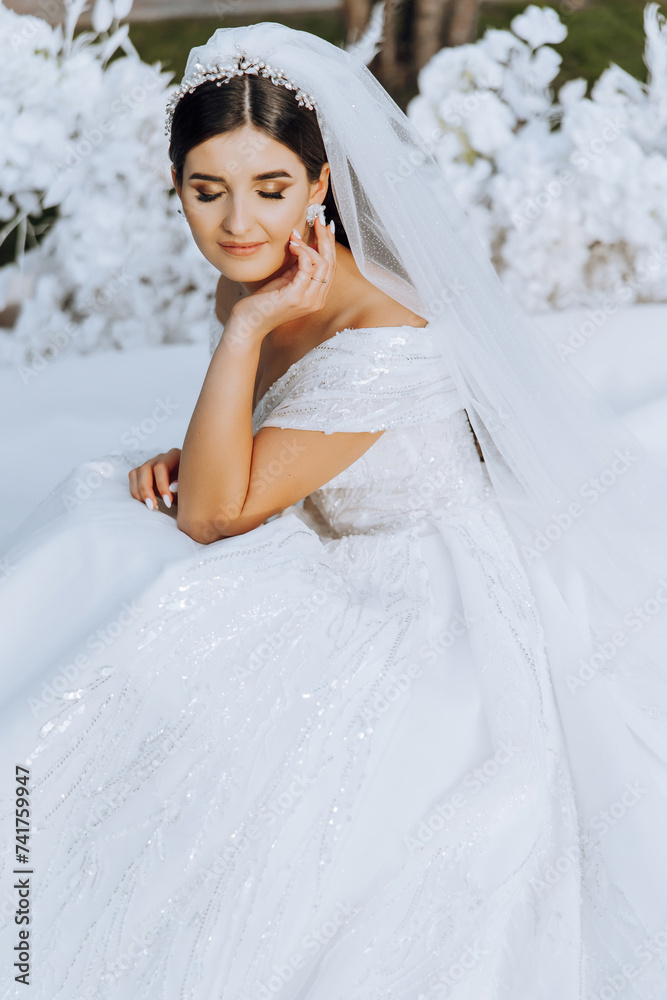 The brunette bride, smiling sincerely, sits on the ground in a unfolded dress, near a white arch made of flowers. Wedding ceremony. Sunny day. Beautiful hair and makeup