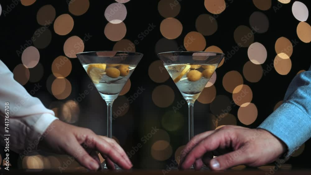 dos copas de cóctel con hielo, limón y aceitunas en un palito. Aparecen dos manos, una mujer con camisa rosa y un hombre con camisa azul, se acarician y luego se alejan. fondo bokeh luces