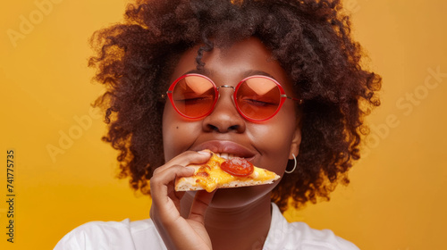 A joyful person is enjoying a slice of pizza, smiling broadly with red glasses, against a yellow background.