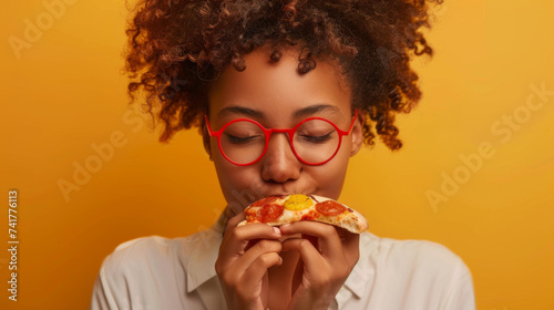 A joyful person is enjoying a slice of pizza, smiling broadly with red glasses, against a yellow background.
