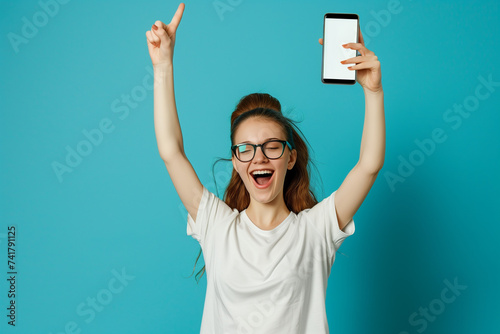 Happy young european lady in casual raises her hand up rejoices online victory and success near huge smartphone with blank screen, isolated on blue background, studio. Win, great app, offer and ad photo