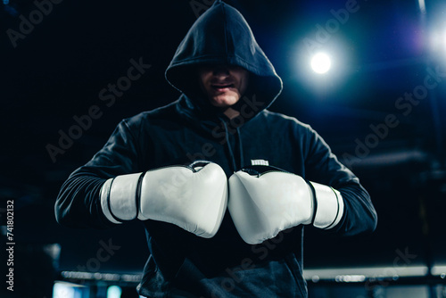 Man in black hoodie is standing in the gym and posing with boxing gloves.